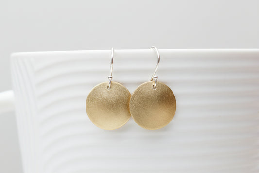A pair of round gold-tone dangle earrings with silver fishhook wires, displayed on the rim of a white textured ceramic cup with a blurred background.