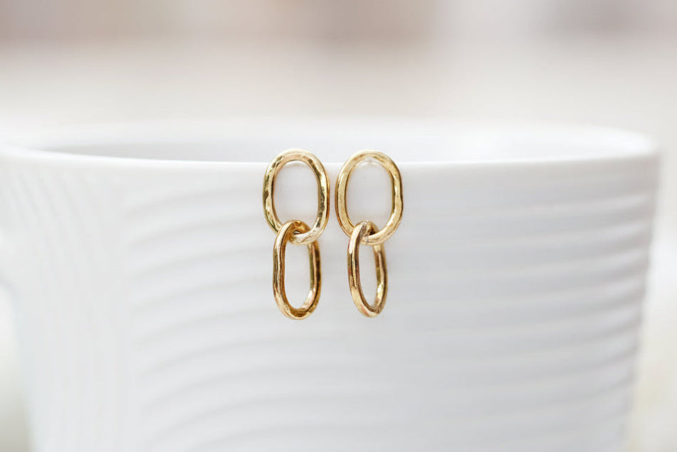A pair of gold chain-link drop earrings resting on the rim of a white textured ceramic mug against a soft, blurred background.