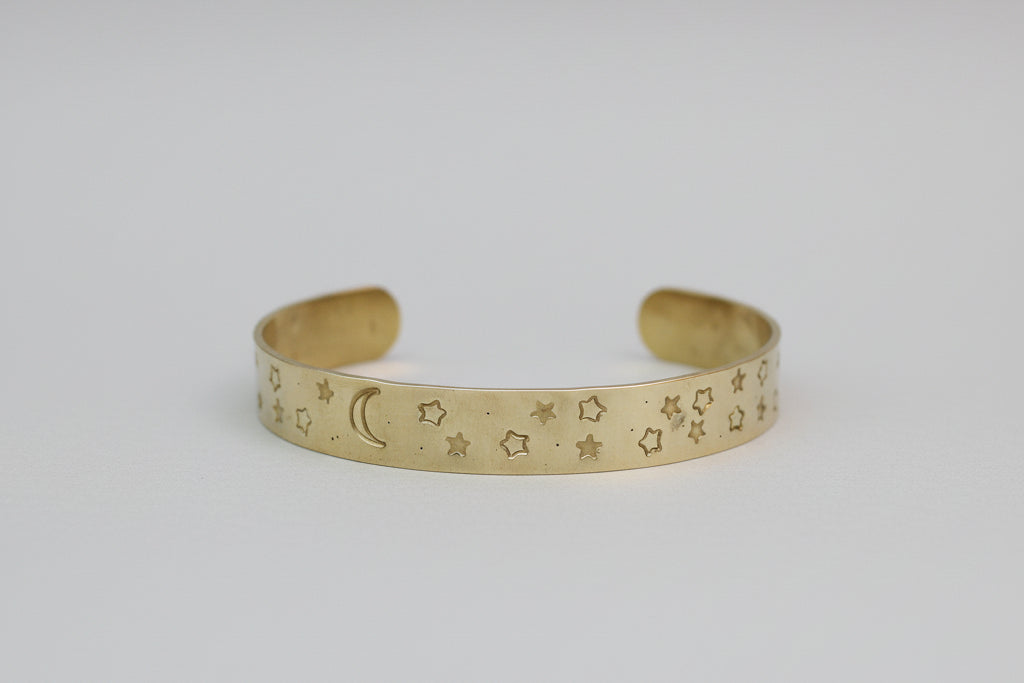 A gold-toned cuff bracelet with engraved star and crescent moon patterns, displayed on a neutral light gray background.