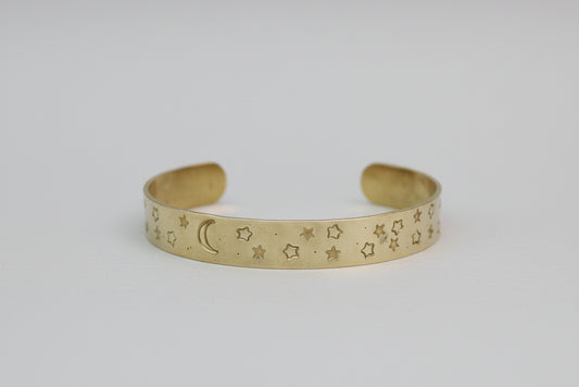 A gold-toned cuff bracelet with engraved star and crescent moon patterns, displayed on a neutral light gray background.