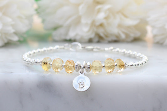 A sterling silver bracelet with citrine gemstones, featuring a disc with an engraved letter 'B' and a small bell-shaped charm.