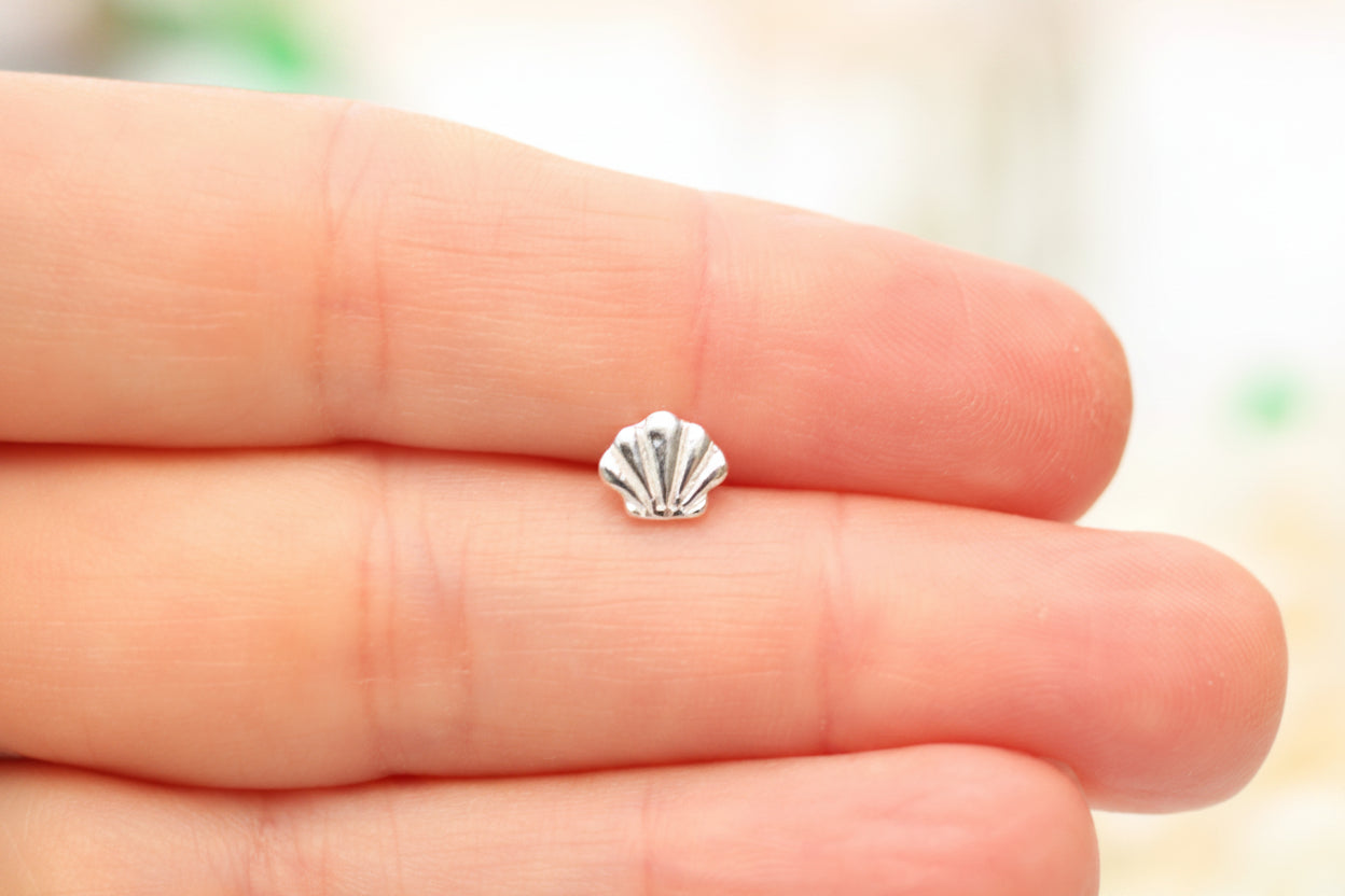 A small silver shell-shaped jewelry charm resting on a person's fingers against a soft, blurred light background.