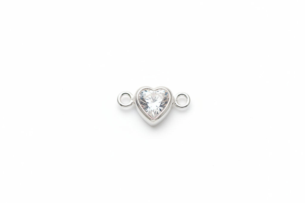 A small silver heart-shaped charm with a clear faceted gemstone in the center, flanked by two small silver loops, resting on a light gray marble surface.
