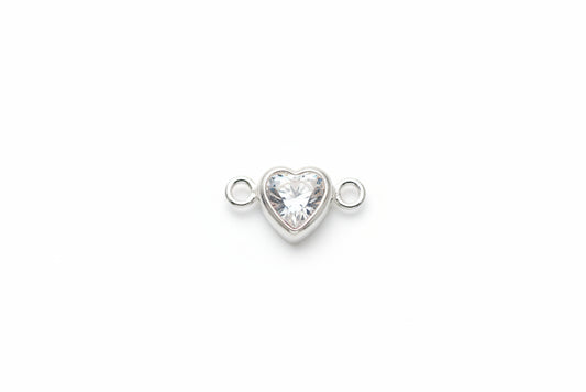 A small silver heart-shaped charm with a clear faceted gemstone in the center, flanked by two small silver loops, resting on a light gray marble surface.