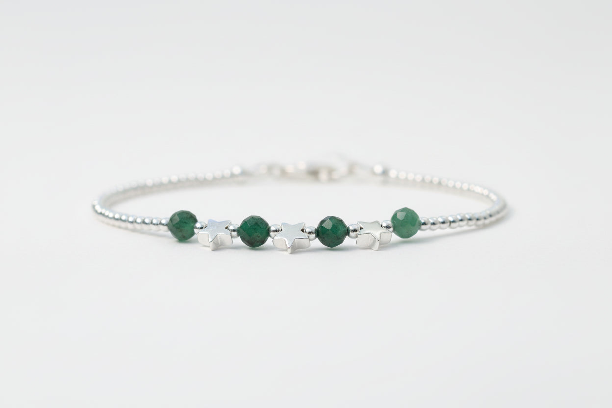 A silver star bracelet with emerald gemstones on a marble surface, with a blurred background of white flowers.