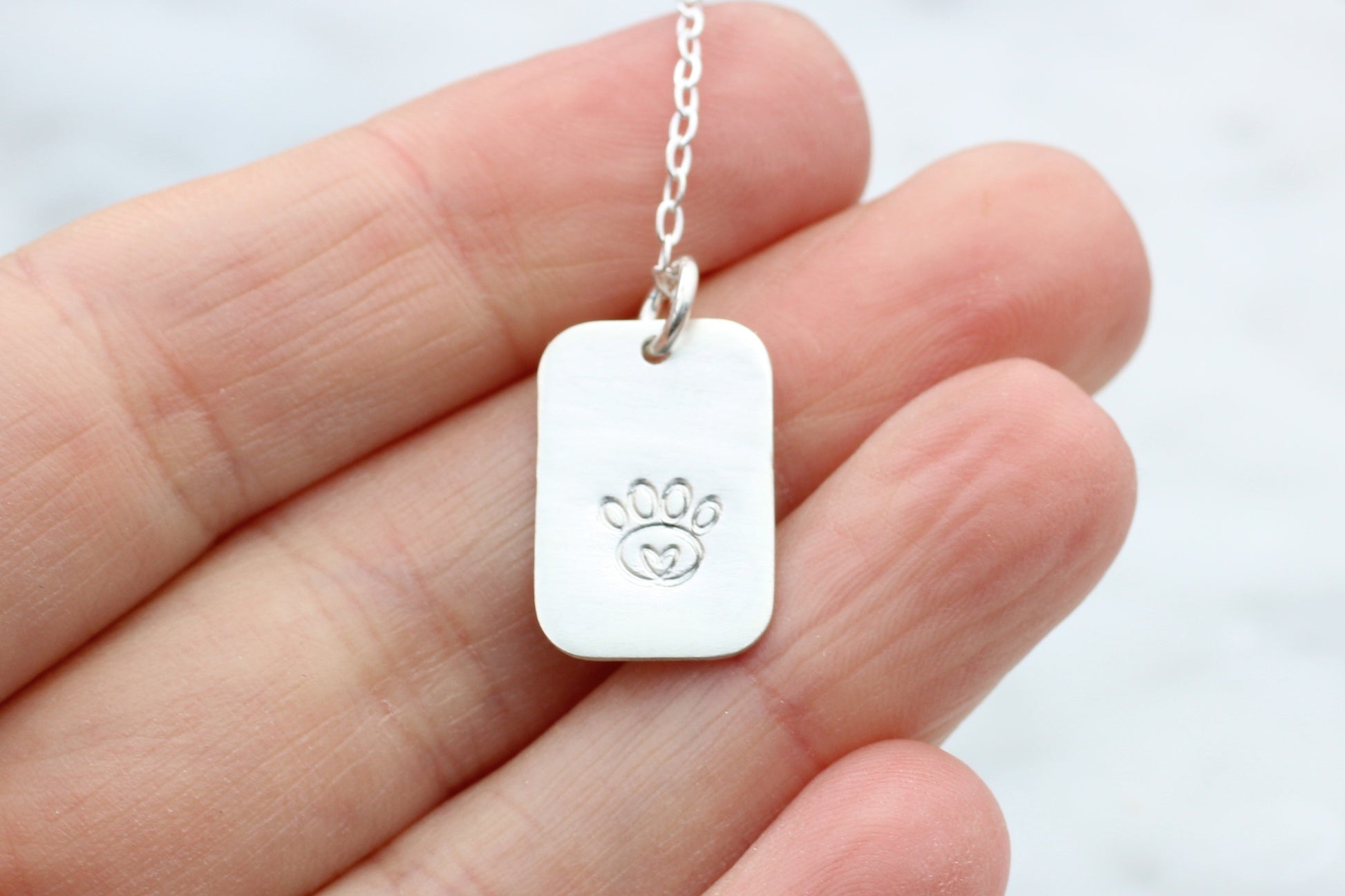 A sterling silver dog tag necklace with a hand-stamped paw print design, held in a person's hand.