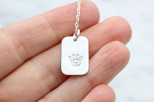 A sterling silver dog tag necklace with a hand-stamped paw print design, held in a person's hand.