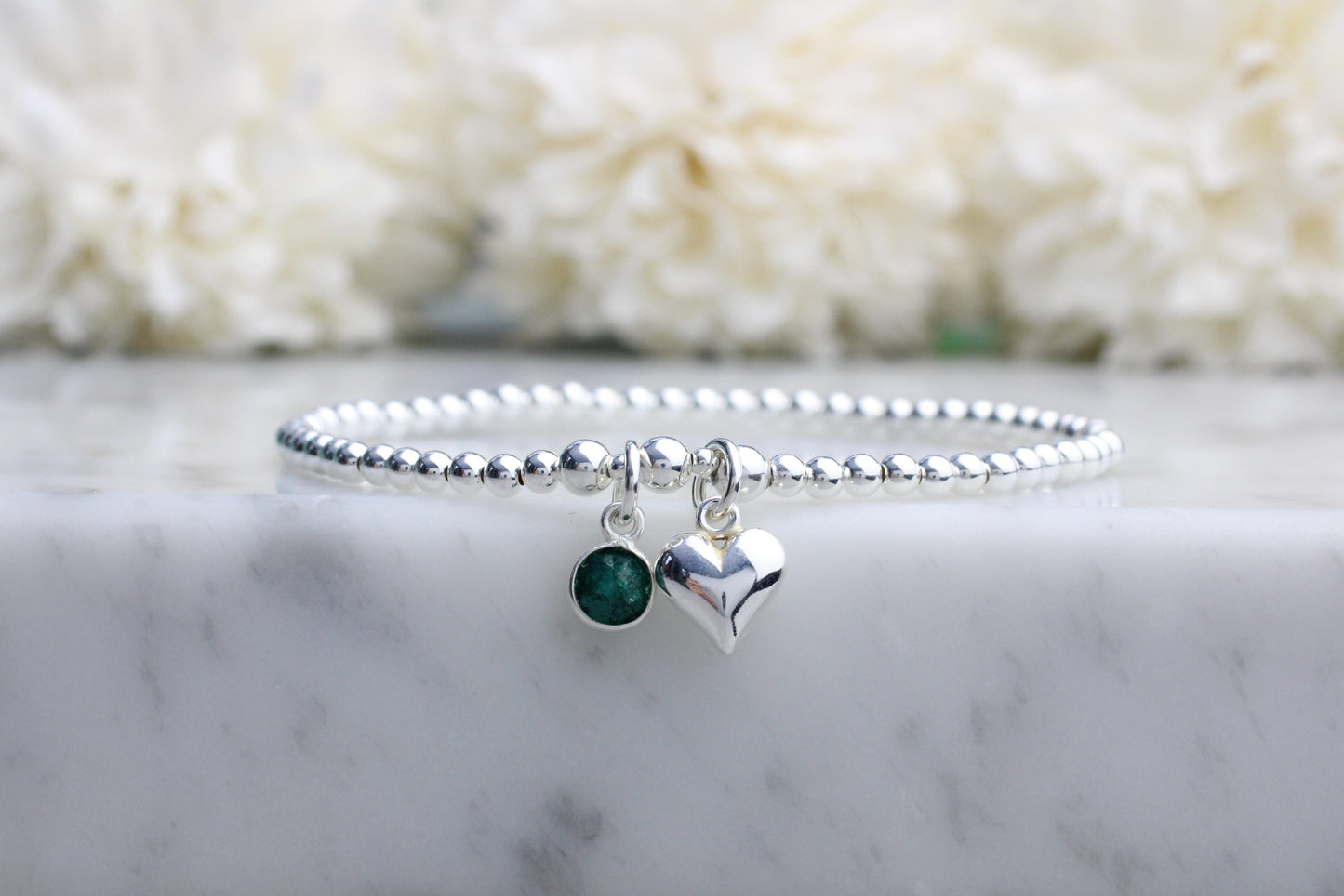 A sterling silver bracelet with a beaded design featuring an emerald green gemstone charm and a silver heart charm, displayed on a marble surface.
