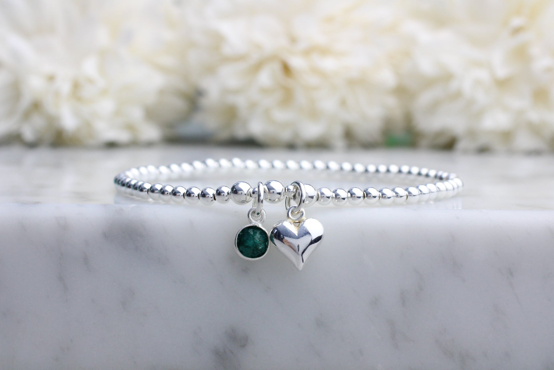 A sterling silver bracelet with a beaded design featuring an emerald green gemstone charm and a silver heart charm, displayed on a marble surface.