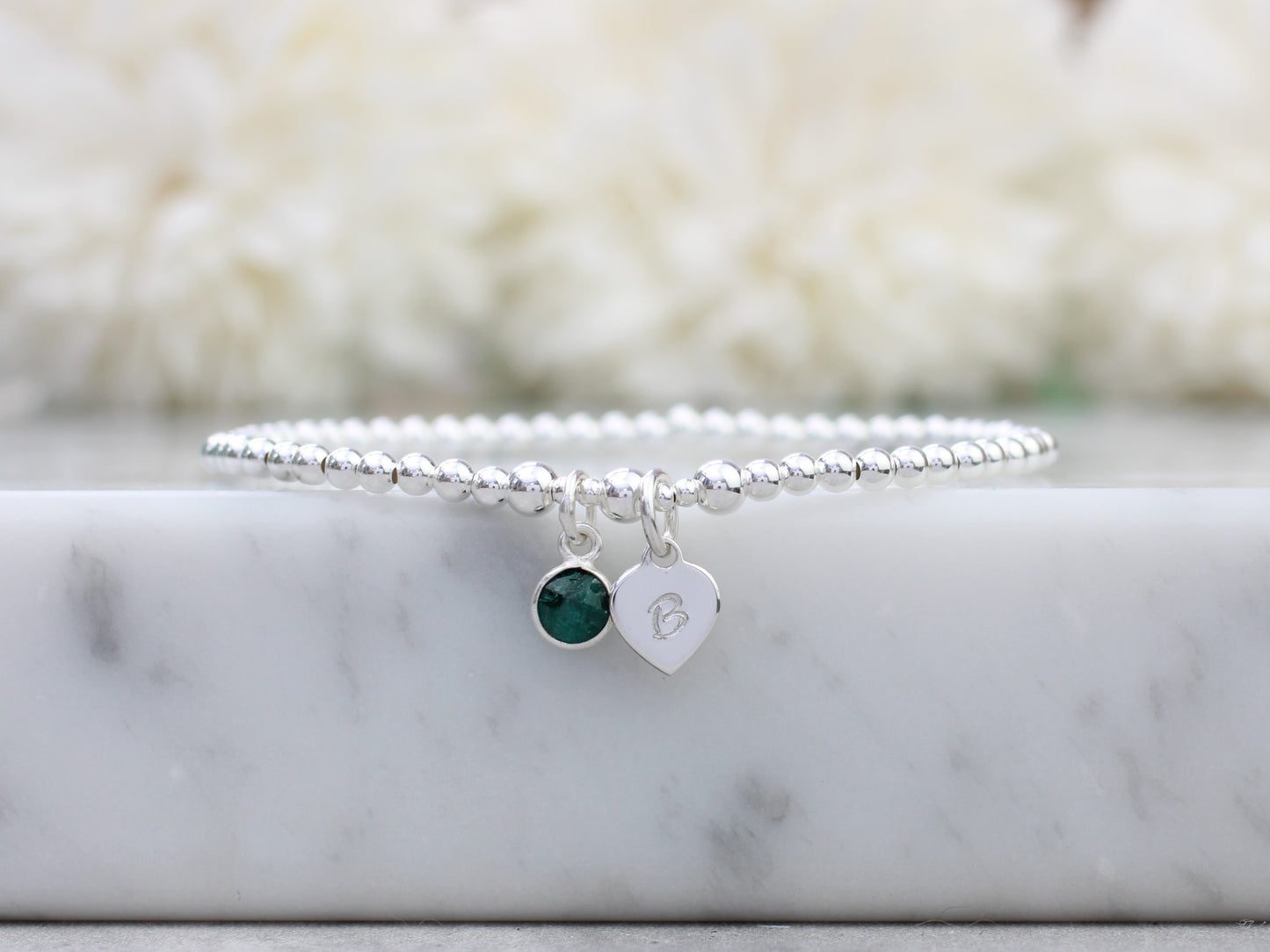 A sterling silver bracelet with a heart charm and an emerald charm, displayed on a marble surface.