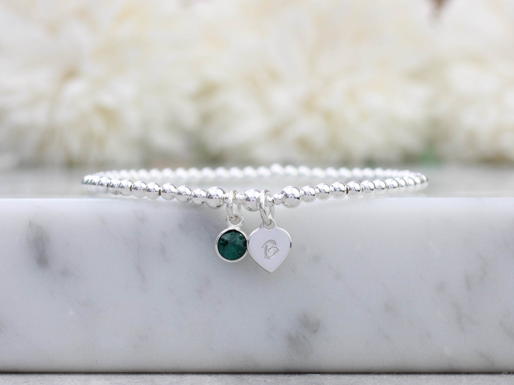 A sterling silver bracelet with a heart charm and an emerald charm, displayed on a marble surface.