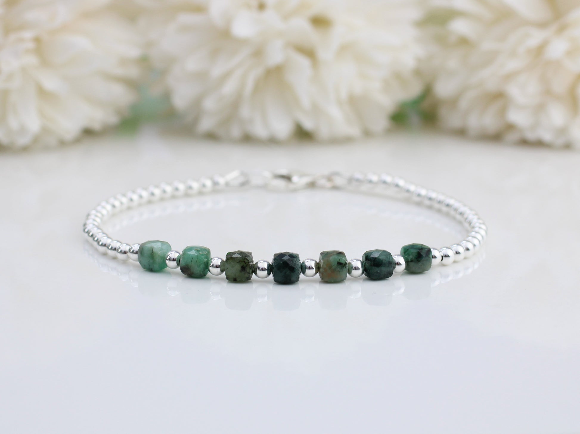 A silver bracelet with multiple green emerald cube gemstones, displayed against a floral background.