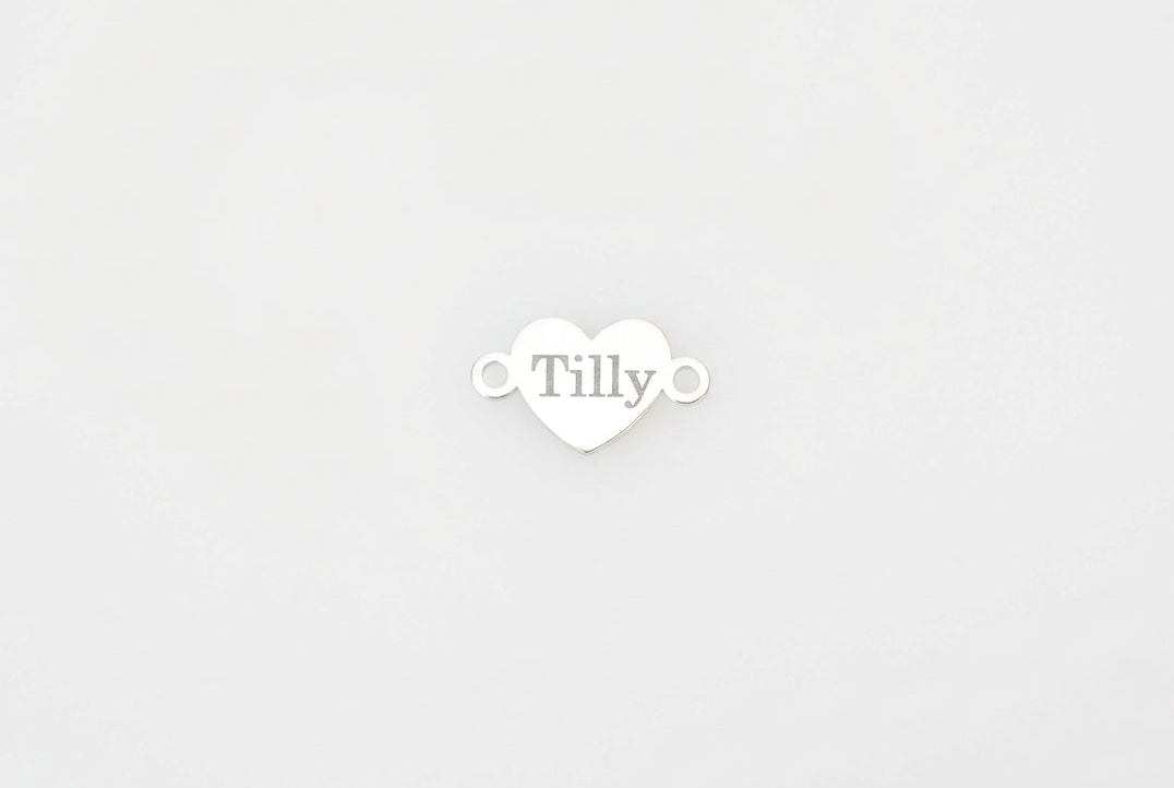 A small white heart-shaped charm with the name 'Tilly' printed in the center, resting on a white marble surface with gray veining.