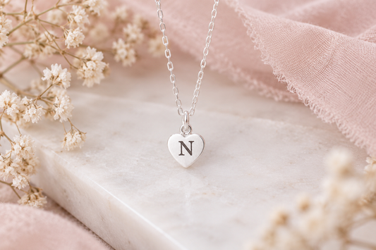 A silver heart pendant necklace with the letter 'N' engraved on it, resting on a white marble slab with soft pink fabric and dried white flowers.