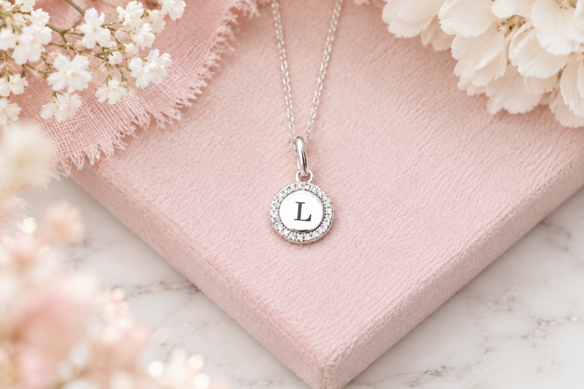 A silver initial pendant necklace with the letter 'L' set in a halo of small clear stones, displayed on a textured pink gift box with white flowers.