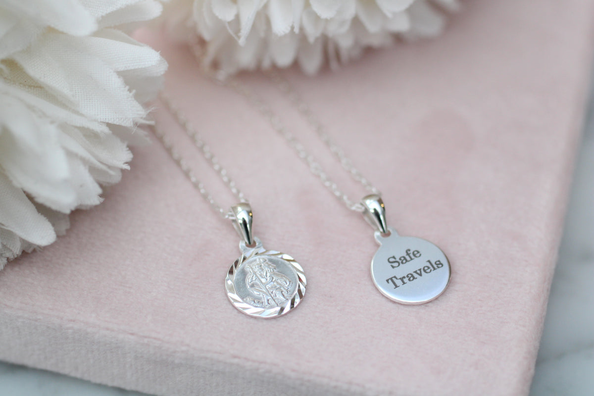 Two silver round pendant necklaces on delicate chains, displayed on a soft pink fabric surface with white flowers. One pendant is engraved with a profile portrait and the other is engraved with the words 'Safe Travels'.