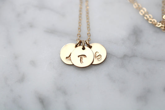 A gold chain necklace with three round disc charms engraved with letters, resting on a white marble surface.