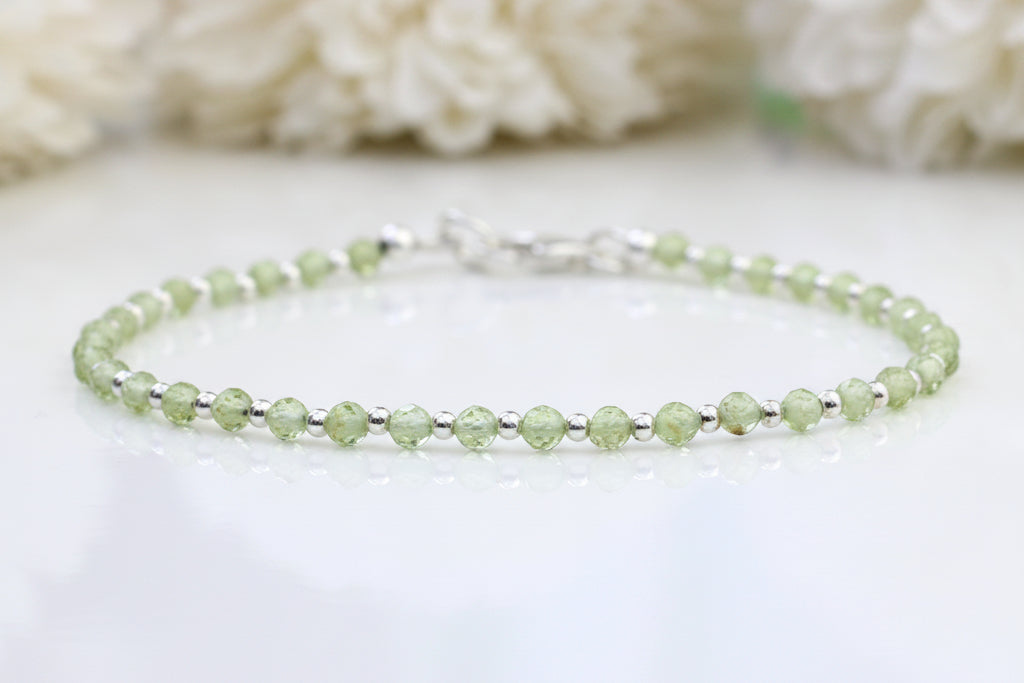 A delicate beaded bracelet made of light green faceted beads separated by small silver spacer beads, with a silver clasp, displayed on a white surface with a soft-focus background of white flowers.