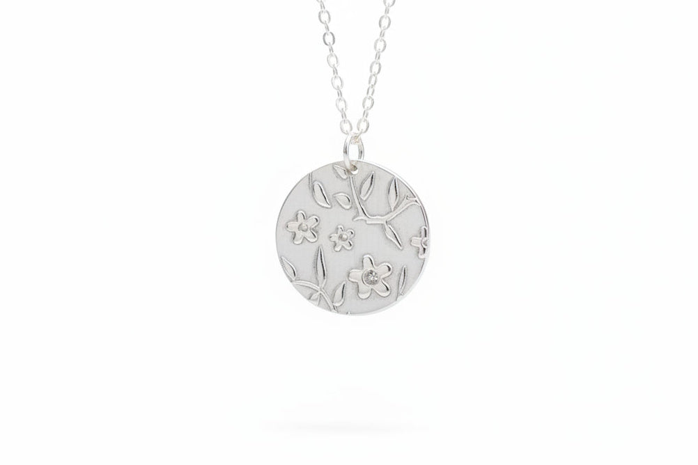 Silver necklace with a round pendant featuring floral designs on a marble surface with white flowers in the background.