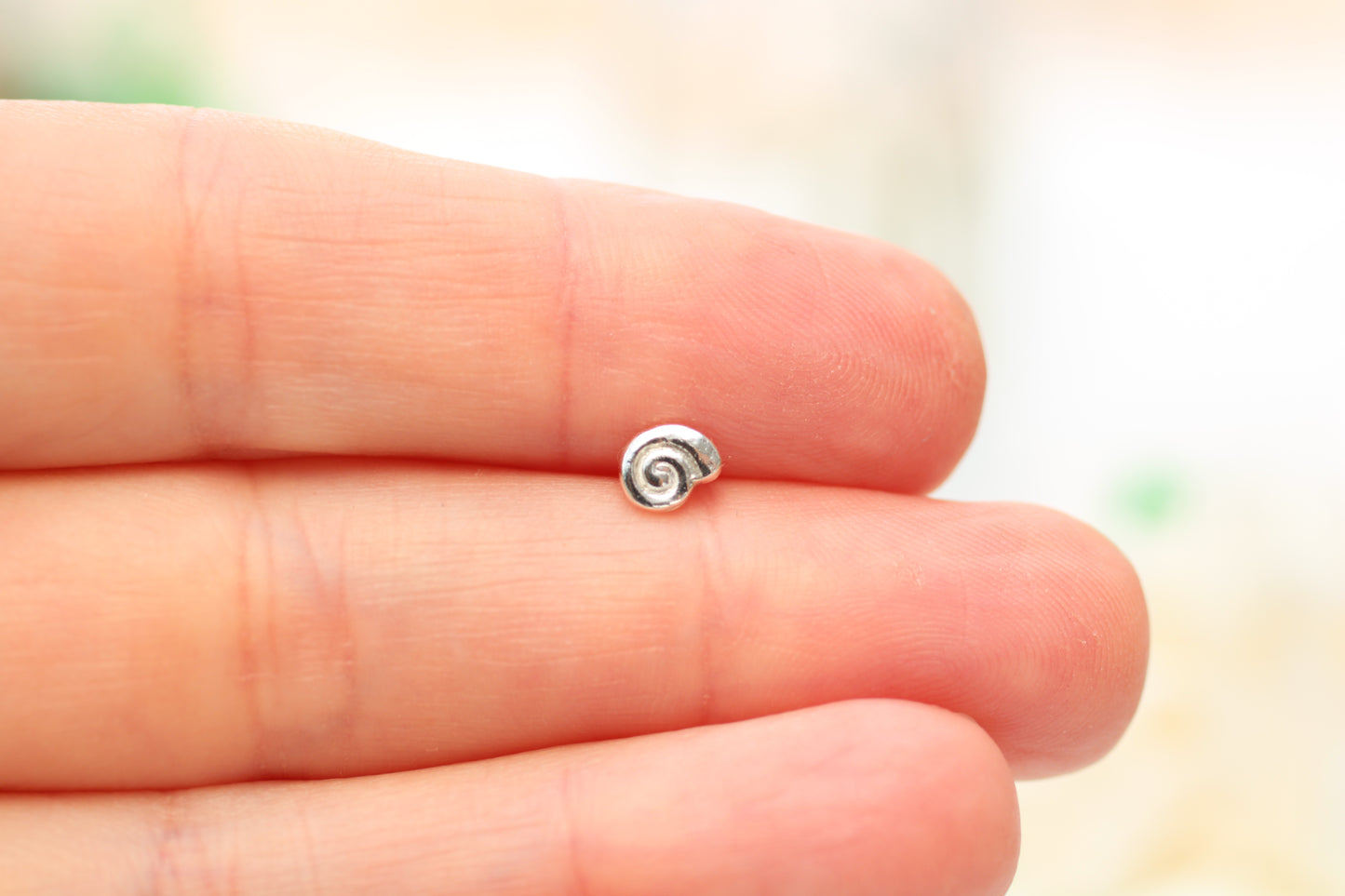 A tiny spiral-shaped silver charm or bead resting on the fingertips of a hand, shown in a close-up shot against a soft, blurred light background.