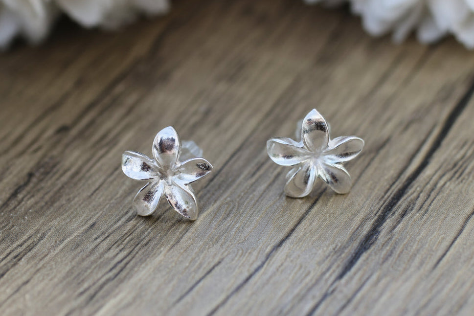 Silver flower-shaped earrings on a wooden surface with white flowers in the background.