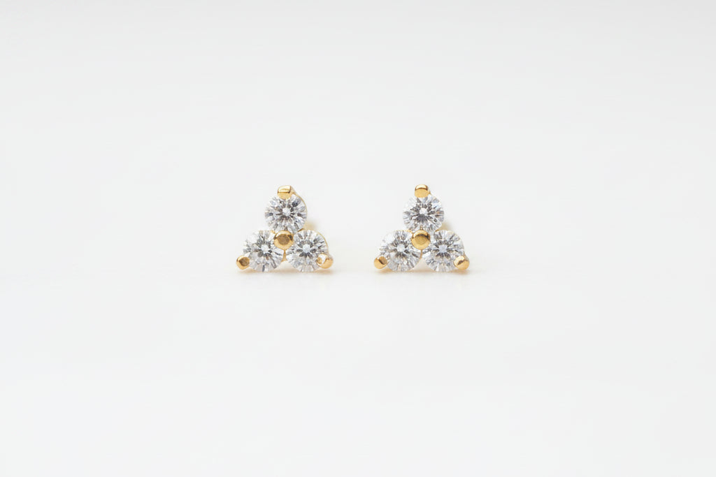 A pair of small triangular stud earrings with clear gemstones and gold-tone accents, resting on a light marble surface.