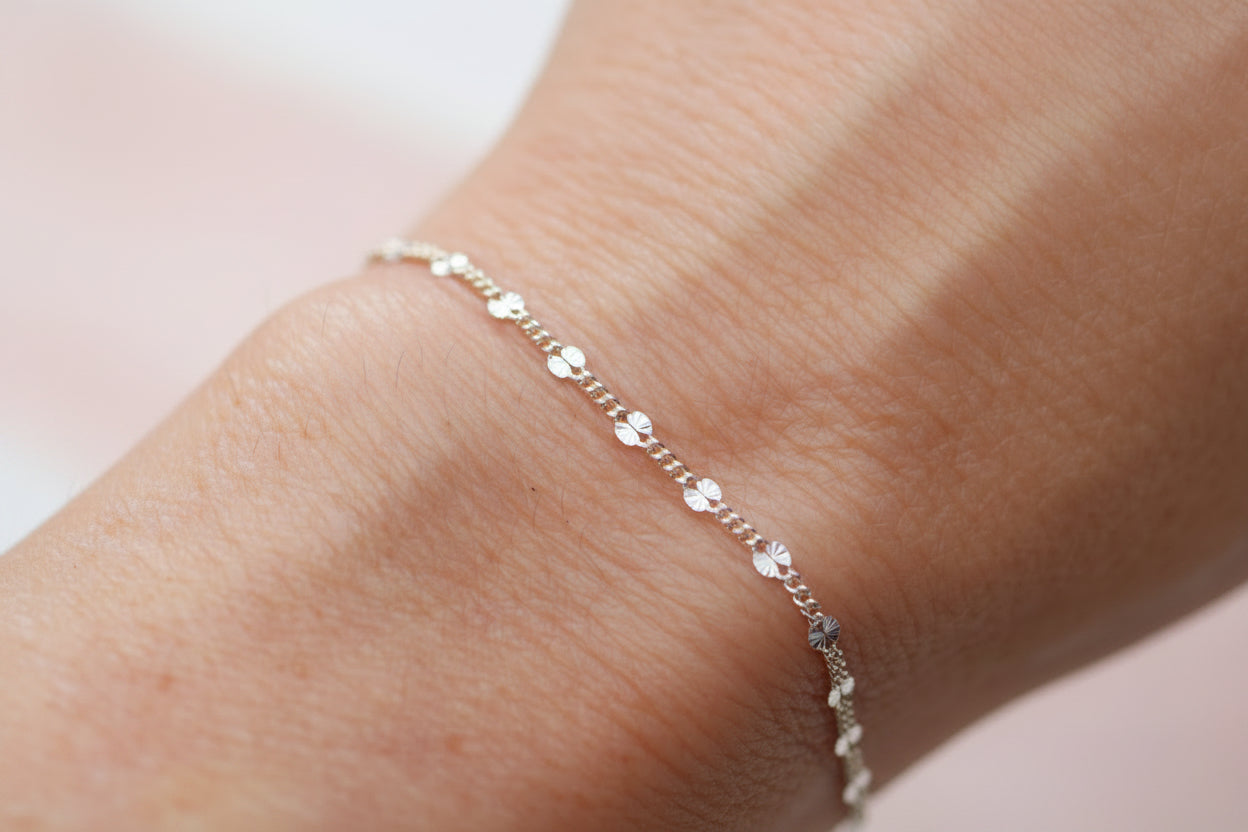A delicate gold chain bracelet with small clear crystal charms, shown worn on a person's wrist against a soft, out-of-focus light gray background.