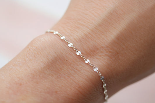 A delicate gold chain bracelet with small clear crystal charms, shown worn on a person's wrist against a soft, out-of-focus light gray background.