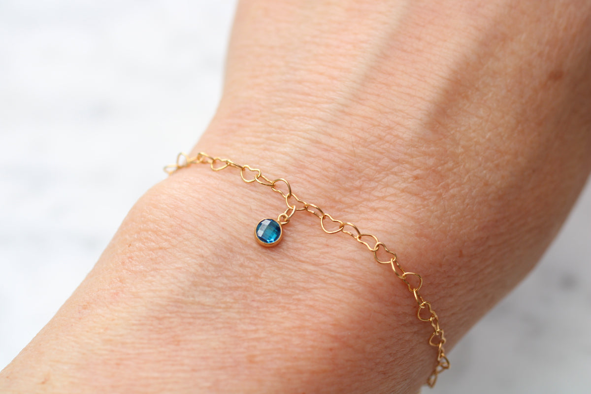 A delicate gold chain bracelet with a small oval blue gemstone charm, shown worn on a person's wrist against a light marble background.