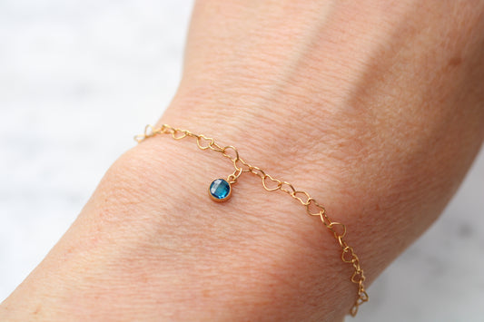 A delicate gold chain bracelet with a small oval blue gemstone charm, shown worn on a person's wrist against a light marble background.