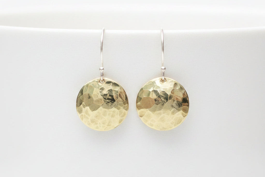 A pair of hammered round gold drop earrings hanging from silver fishhook wires, displayed against a white textured ceramic mug in a softly blurred indoor setting.