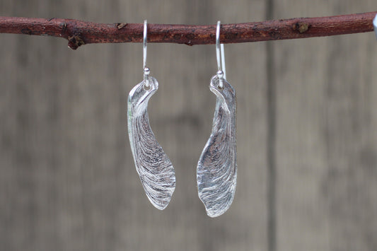 A pair of silver-toned dangle earrings with organic, leaf-like translucent shapes, hanging from a brown twig against a blurred gray-brown wooden background.