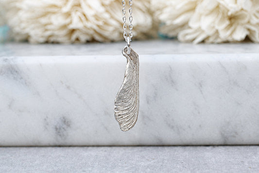 A silver-toned pendant necklace with a textured, organic leaf or feather-like shape, hanging against a soft-focus background of a white marble ledge and cream flowers.