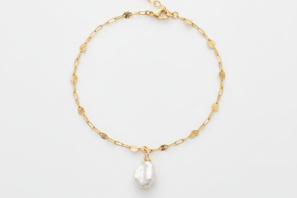 A delicate gold chain bracelet with small gold beads and a teardrop-shaped white pearl pendant, displayed on a white marble surface with white flowers in the background.