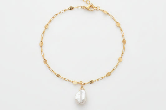 A delicate gold chain bracelet with small gold beads and a teardrop-shaped white pearl pendant, displayed on a white marble surface with white flowers in the background.