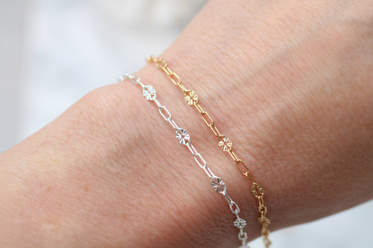 Two delicate chain bracelets on a person's wrist, one in silver with star and flower motifs, and one in yellow gold with matching floral links.