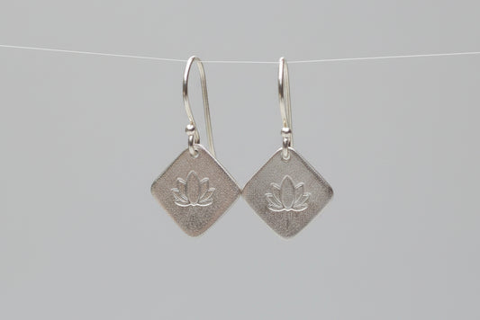 A pair of silver drop earrings with textured square pendants featuring an embossed lotus flower, hanging from silver fishhook wires from the rim of a white ceramic cup.