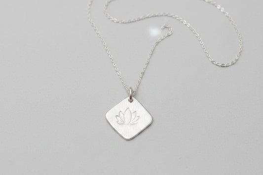 A silver necklace with a square pendant featuring an engraved lotus flower, displayed on a white and gray marble surface.
