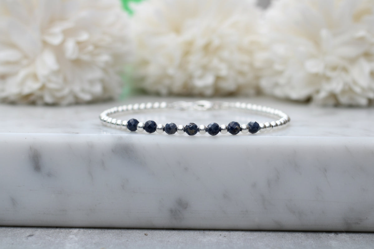 A delicate silver beaded bracelet with five dark navy blue faceted beads, displayed on a white marble slab with soft white flowers in the background.