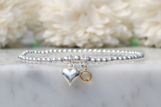 A silver beaded bracelet with a polished heart charm and a small round amber gemstone charm, displayed on a white marble surface with soft white flowers in the background.