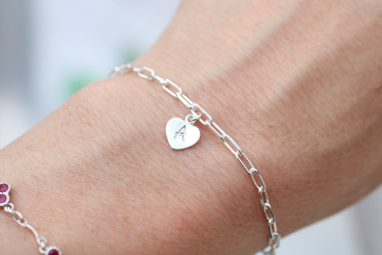 A close-up of a delicate silver chain bracelet with a small heart-shaped charm engraved with the letter 'A', worn on a person's wrist.