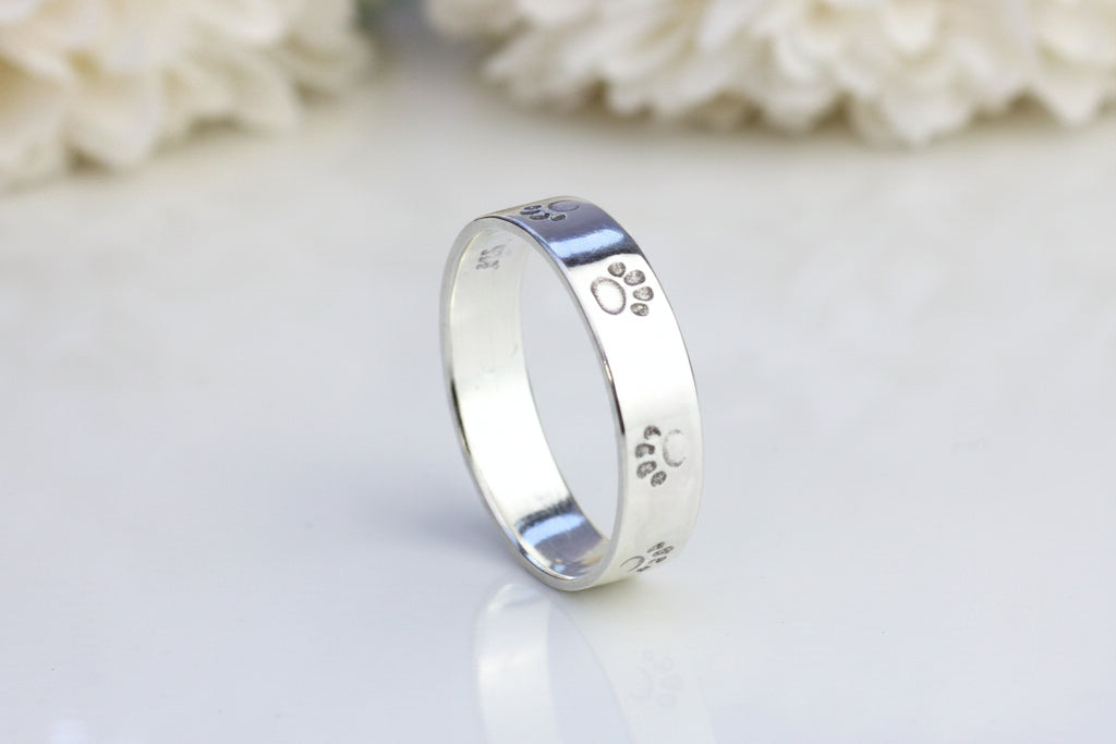 A silver paw print ring on a reflective surface, with a blurred background of flowers.
