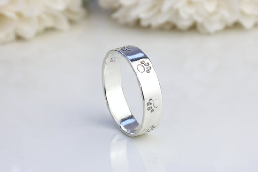 A silver paw print ring on a reflective surface, with a blurred background of flowers.