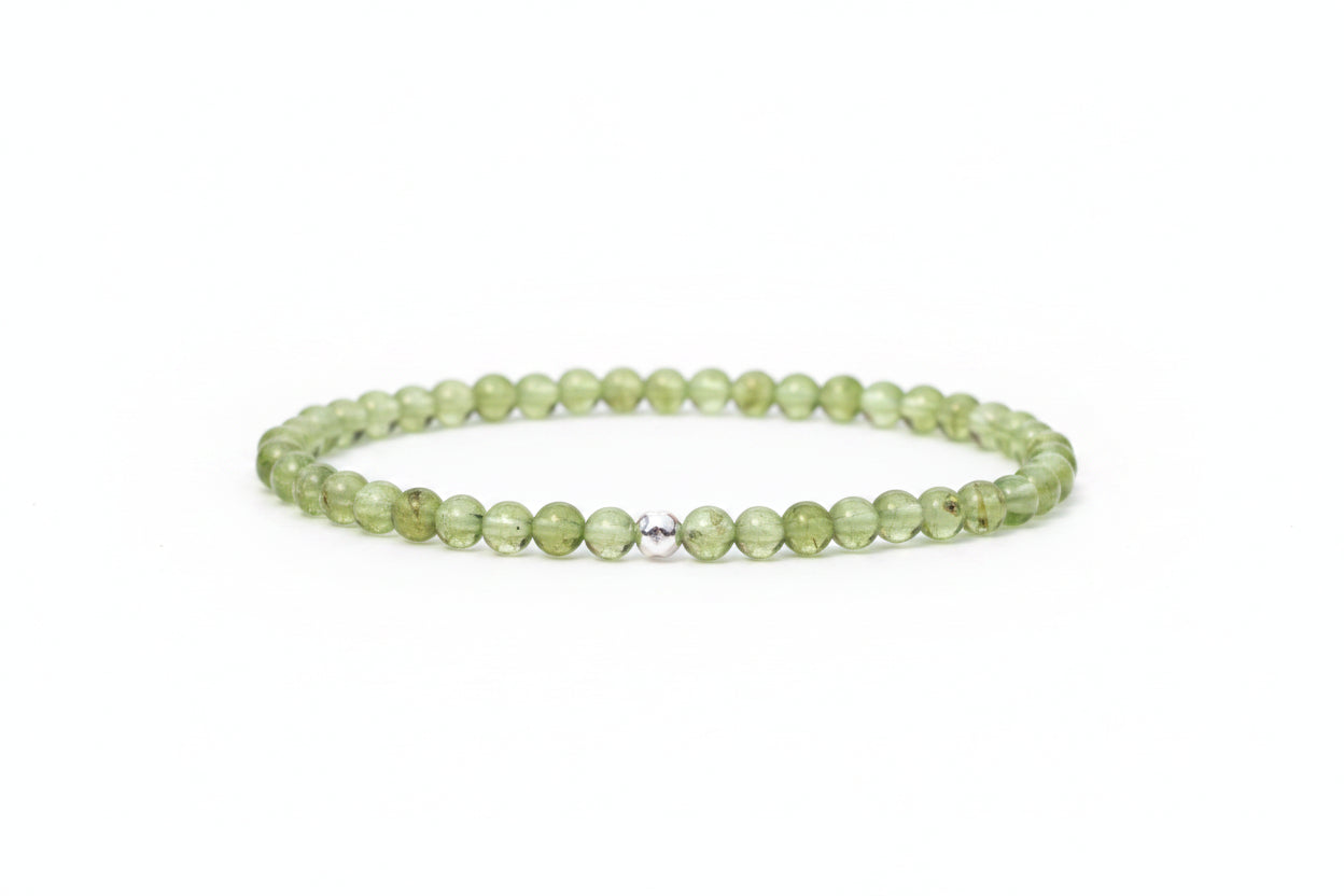 A beaded stretch bracelet made of light green faceted beads with a small silver-toned metal accent bead in the center, resting on a white marble surface with blurred white flowers in the background.