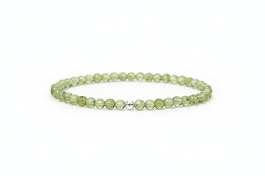 A beaded stretch bracelet made of light green faceted beads with a small silver-toned metal accent bead in the center, resting on a white marble surface with blurred white flowers in the background.