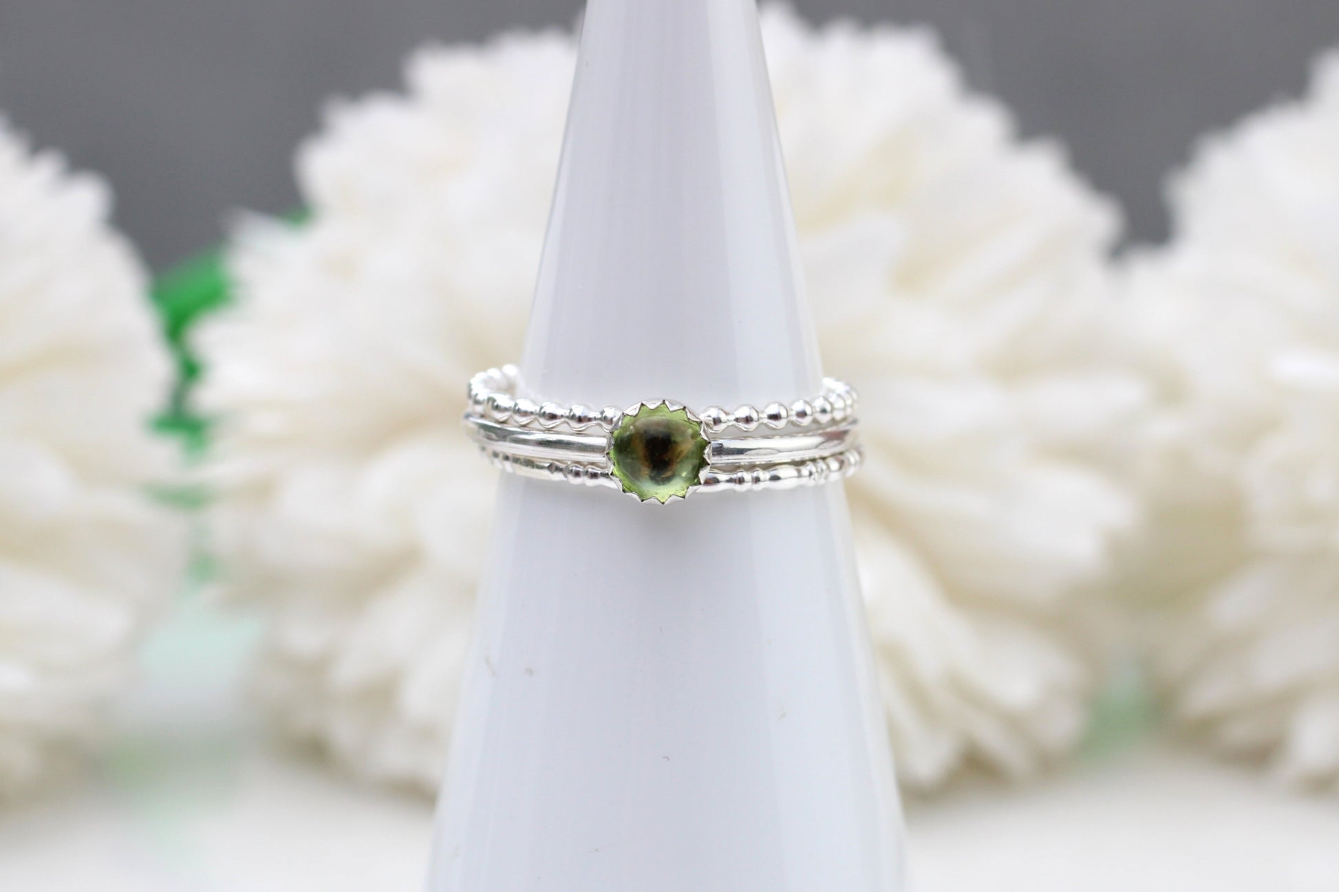 Silver ring with a green gemstone on a white stand against a blurred floral background