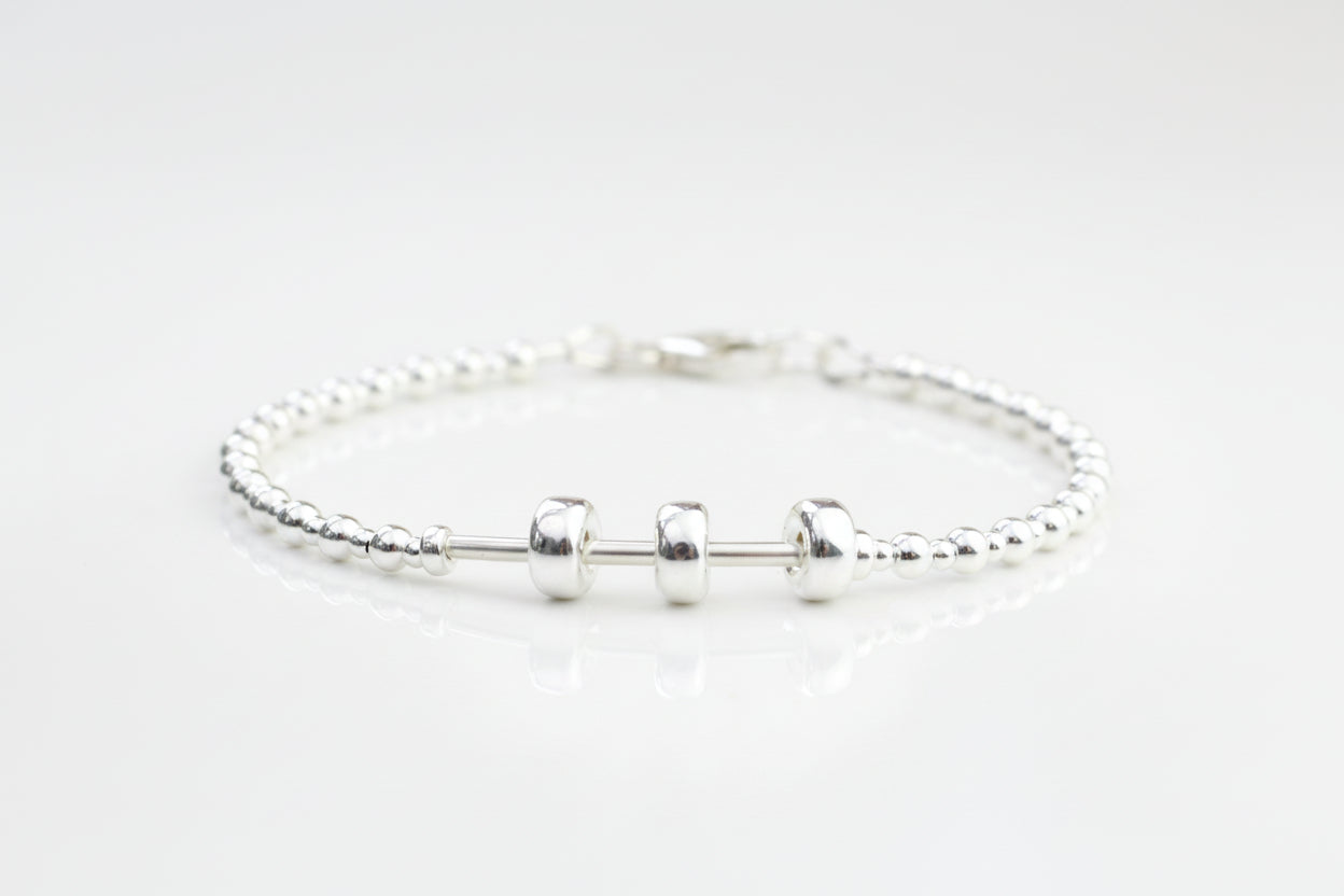 A sterling silver bracelet with three moving beads on a textured silver band, presented on a white surface with flowers in the background.