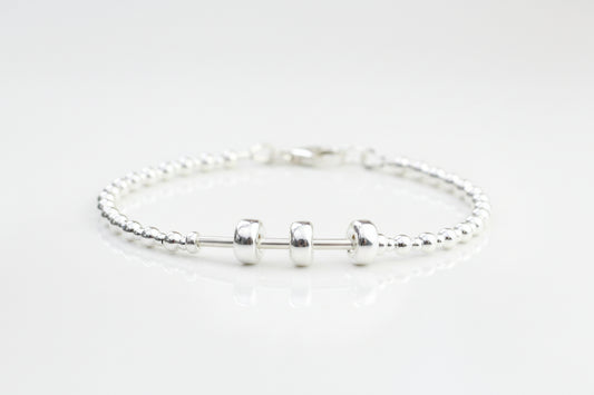 A sterling silver bracelet with three moving beads on a textured silver band, presented on a white surface with flowers in the background.