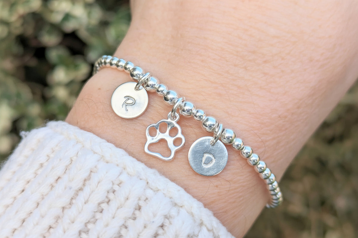 A sterling silver bracelet with a paw print charm and beads, featuring a personalized initial disc.