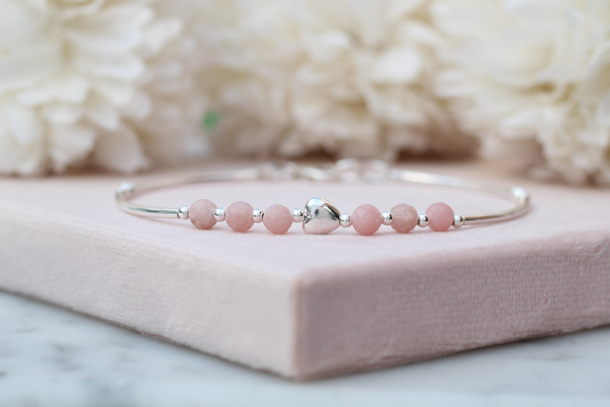 A silver-toned bangle bracelet with seven light pink round beads and a small silver heart charm, displayed on a pink block with soft white flowers in the background.